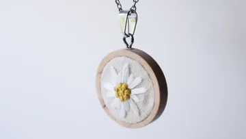 Embroidered daisy pendant on chain, white background.