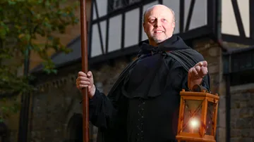 Man in cloak holding lantern outside building.