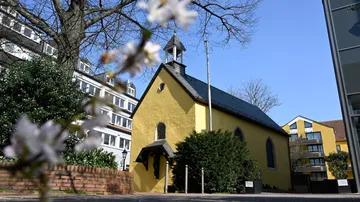 Gelbe Kapelle umgeben von Bäumen und Gebäuden.