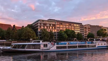 Cruise ship on river at sunset, modern buildings.
