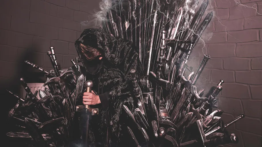 Person in cloak sitting on iron throne indoors.