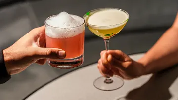 Two hands holding cocktails, toasting indoors.