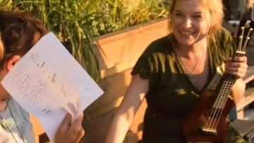 Woman holding ukulele, sitting outdoors, smiling.