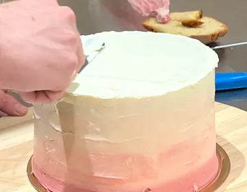 Hände glasieren einen rosa Verlaufskuchen auf Tisch.