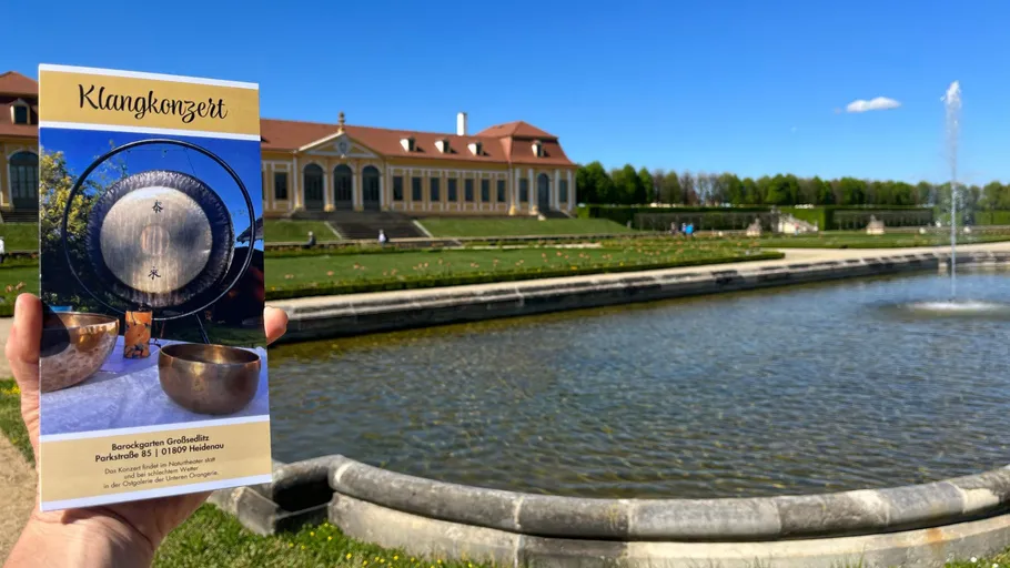 Person hält Flyer vor Gebäude und Brunnen.