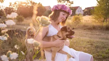 Frau mit Blumenkron hält kleinen Hund im Freien.