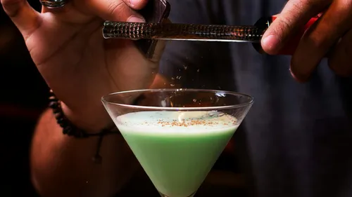 Person grating spice over green cocktail glass.