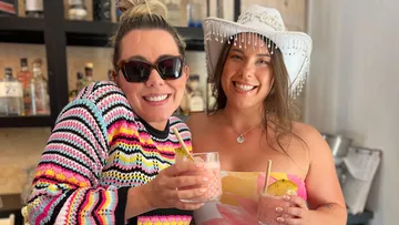 Two women smiling, holding drinks, indoors.