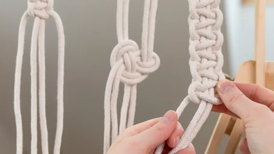 Hands crafting macramé with white rope indoors.