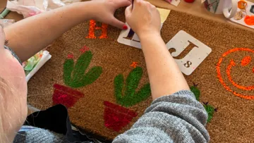 Person painting letters and cacti on doormat.
