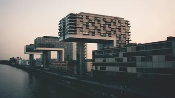 Modern buildings resembling cranes over riverbank.