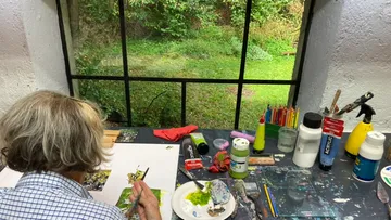 Person painting at table near garden window.