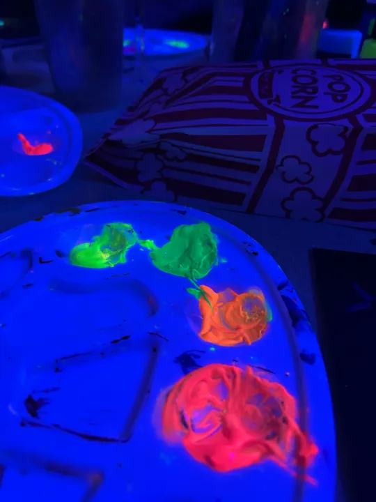 Neon paints glowing under blacklight beside popcorn bag.