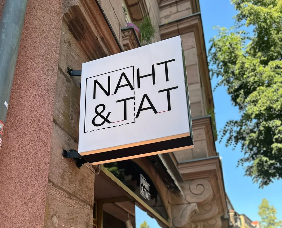 Ladenschild mit 'NAHT & TAT', städtischer Hintergrund.
