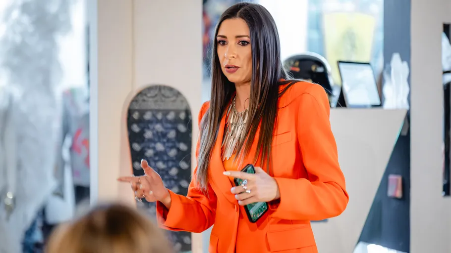 Woman in orange jacket speaking indoors.