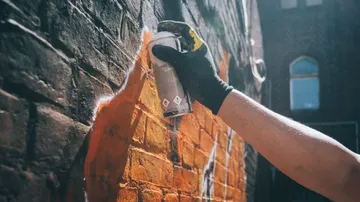 Gloved hand spray-painting orange graffiti on brick wall.