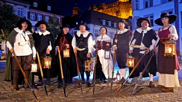 Personen in historischen Kostümen mit Laternen, draußen.