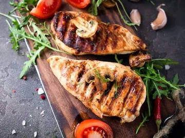 Gegrillte Hähnchenbrust auf Holzbrett mit Gemüse und Gewürzen.