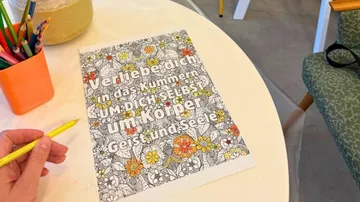 Malbuchseite auf Tisch mit Stiften, Hand hält Stift.