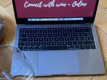 Laptop zeigt online Wein-Event, Glas daneben auf Holzboden.