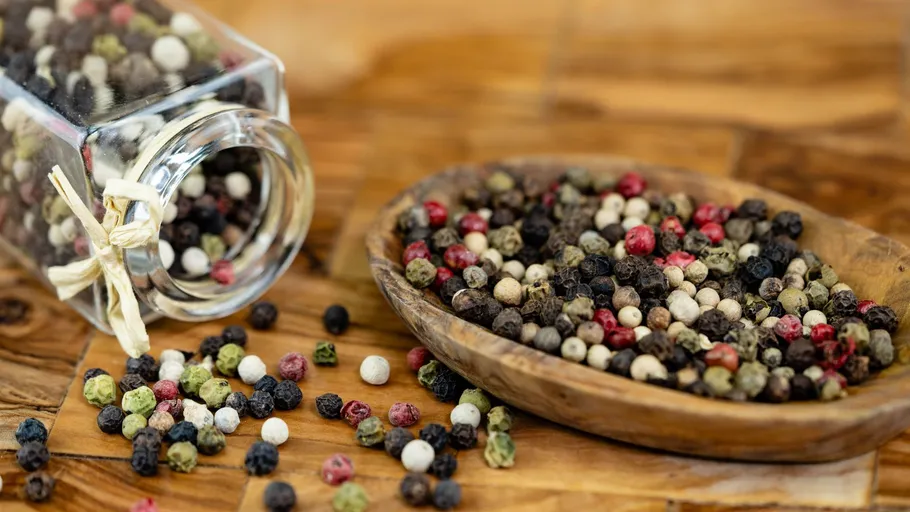 Mixed peppercorns spill from glass jar.