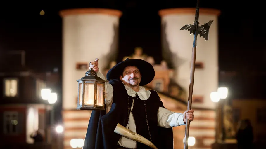 Man dressed as night watchman holds lantern, staff.