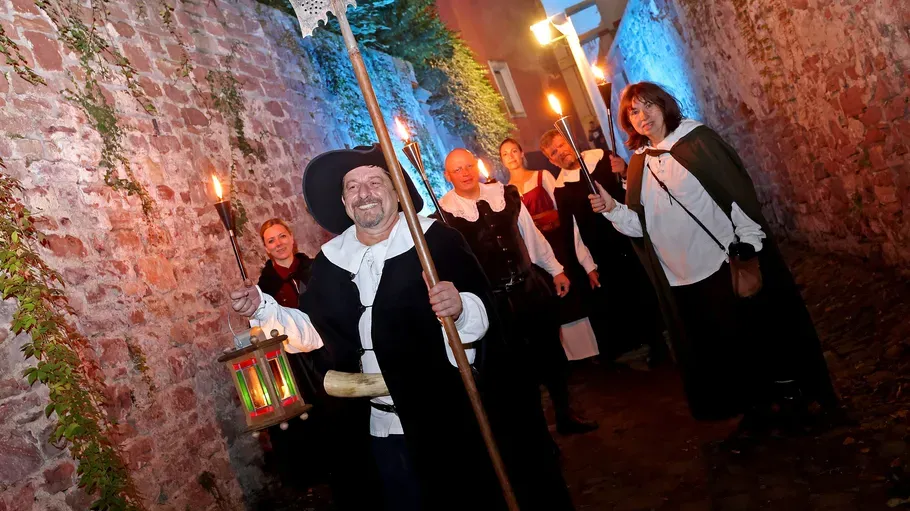 Group in historic clothing holding torches, alleyway setting.