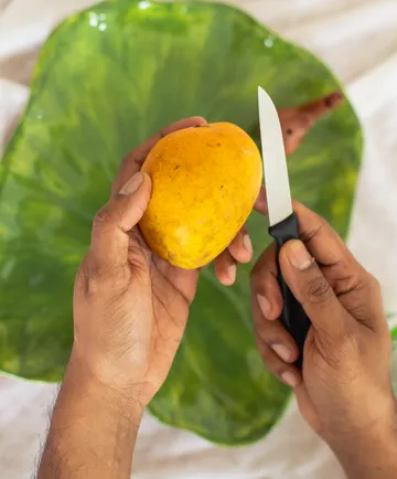 Hände halten Messer und Mango über einem Teller.