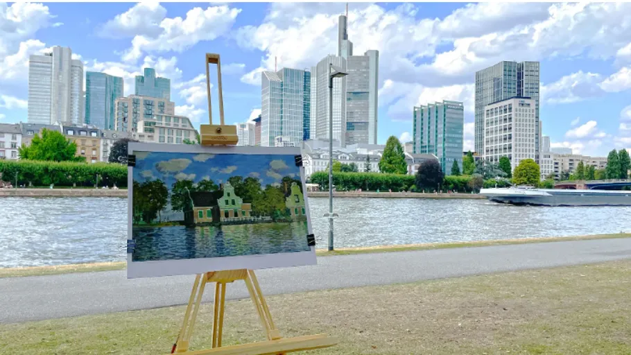 Painting on easel beside river, city skyline background.