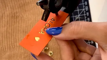 Hand embossing orange tag with "ALOHA" text.