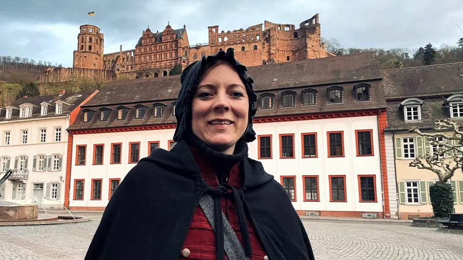 Frau im Umhang lächelt, Schloss im Hintergrund.