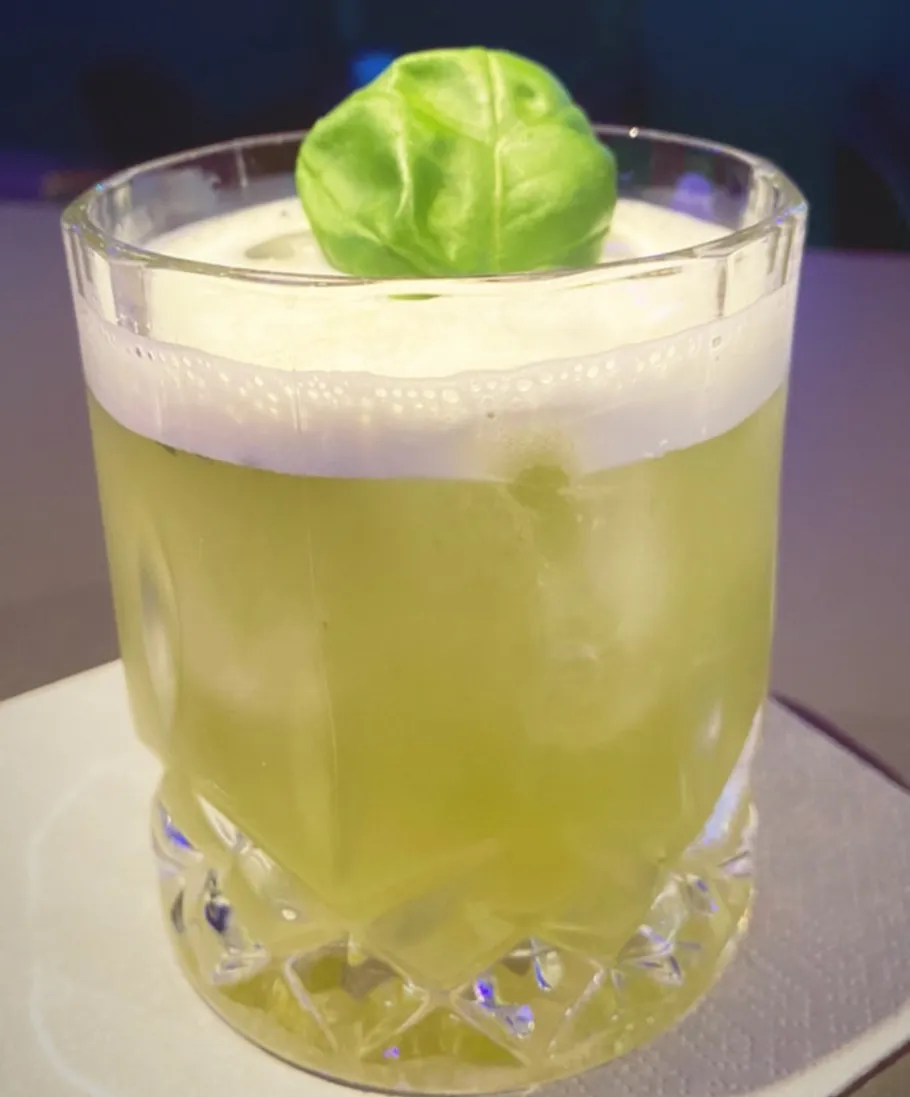 Green cocktail with foam, garnished with basil.