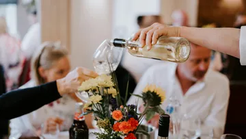 Person gießt Weißwein an einem Tisch ein.