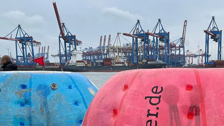 Cranes at port, colorful barrels foreground.