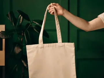 One hand holds a beige tote bag against a green background.