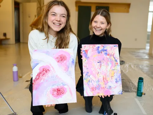 Two women smiling, holding colorful paintings indoors.