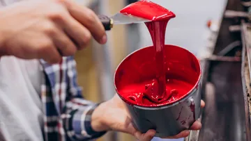 Person stirring red paint with a spatula.