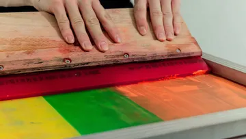 Hands using squeegee on colored screenprint.