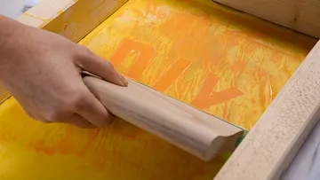Hand using squeegee on yellow screen printing frame.