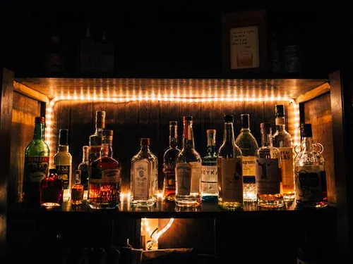 Liquor bottles stand on an illuminated shelf in the dark.