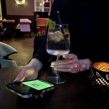 Person hält Getränk und checkt Handy an der Bar.