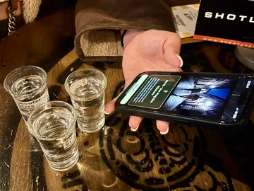 Hand hält Handy, drei Schnapsgläser daneben.