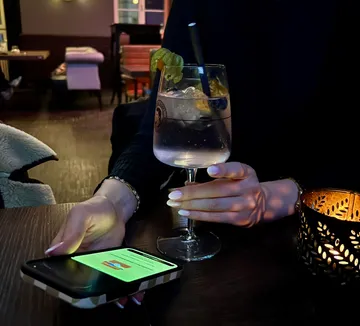 Person hält Getränk, Handy auf Tisch, schwach beleuchtetes Restaurant.