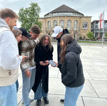 Gruppe lacht über Handys vor Theatergebäude.