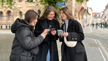 Drei Frauen lachen beim Blick auf Handys draußen.