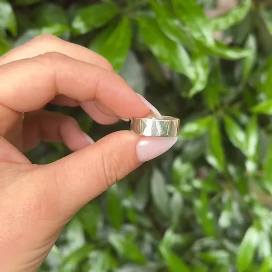 Hand hält einen goldenen Ring, grüner Blätterhintergrund.