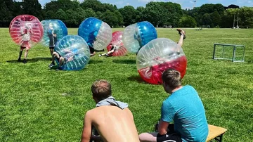 People playing bubble soccer on grass field.