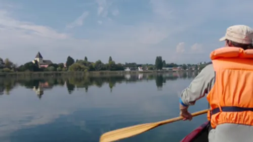 Person im Kajak auf ruhigem See, Dorf sichtbar.