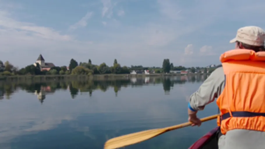 Person im Kajak auf ruhigem See, Dorf sichtbar.