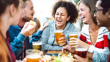 Group laughing, holding beers, outdoor setting.